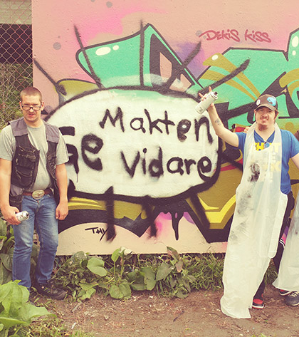 Två män står på varsin sida om en vägg där det står skrivet "Ge makten vidare" med sprayfärg.
