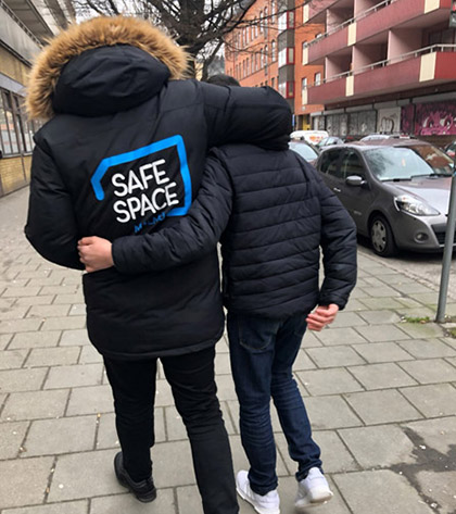 Två personer bakifrån som håller armarna om varandras ryggar medan de går. Texten Safe space står på den ena personens jacka.
