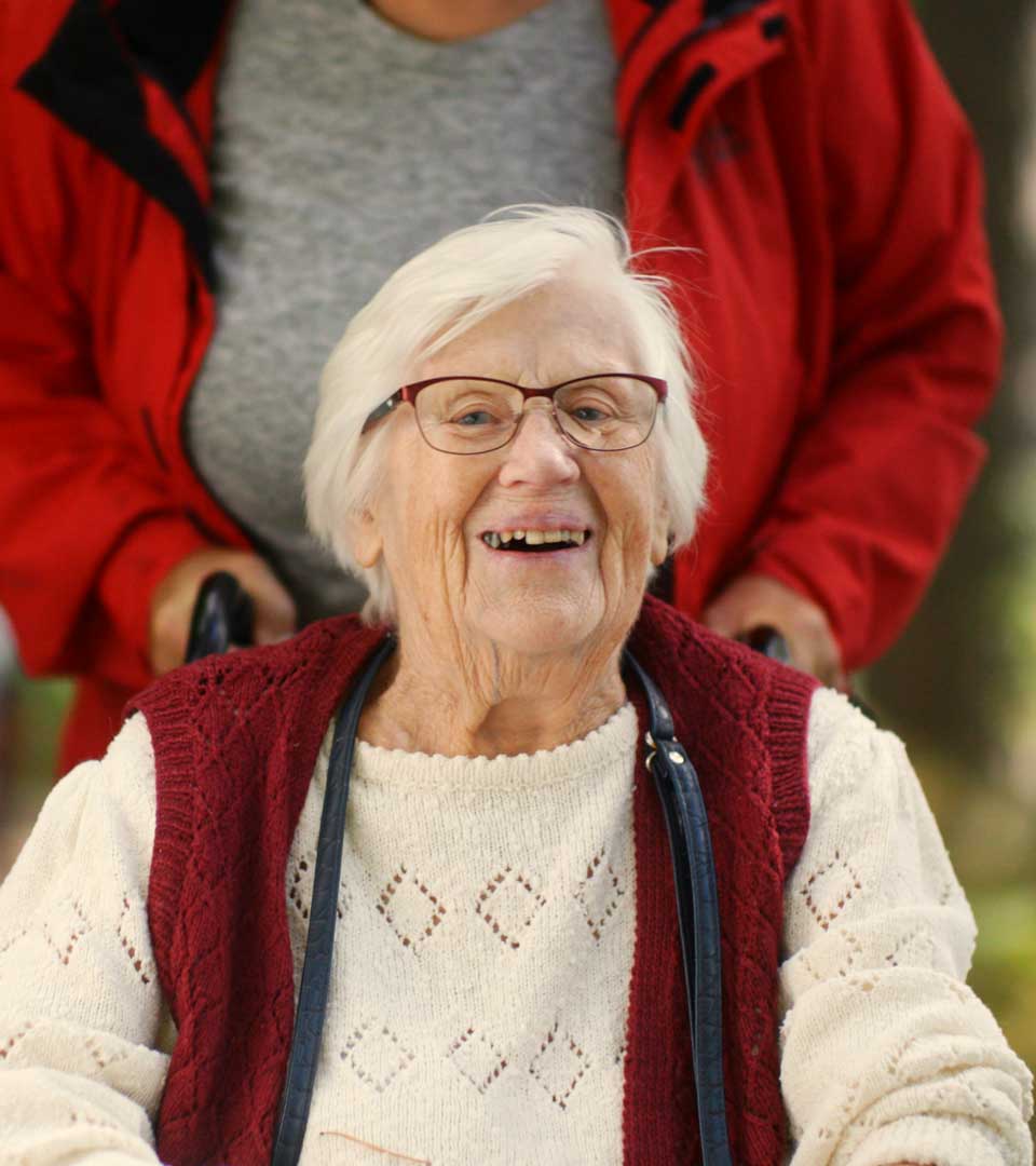 En äldre kvinna i rullstol ler och tittar in i kameran.