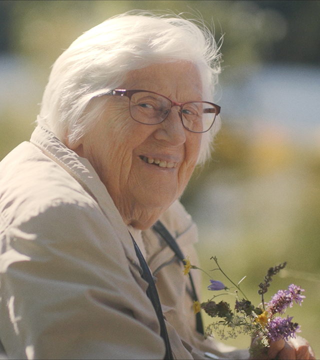 Porträttfoto av en äldre kvinna som leende tittar in i kameran. Hon bär en beige jacka och håller en bukett vilda blommor i händerna. 