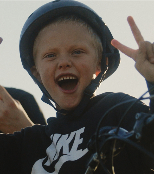 Én pojke som bär hjälm håller upp fingrarna i ett segertecken. Han har munnen öppen och ser ut att vara glad.