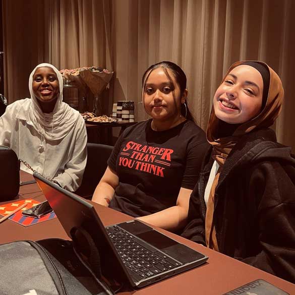 Tre tjejer sitter framför ett bord med en dator. Alla tittar in i kameran och ser glada ut.