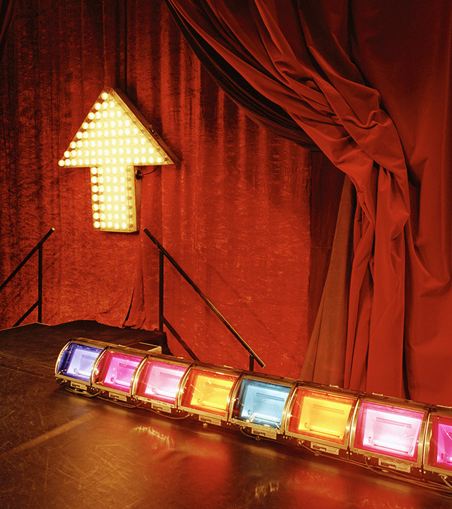 På ett scengolv står en rad med färgglada strålkastare.På väggen bakom scenen hänger ett rött draperi. På väggen hänger en pil som pekar uppåt. Pilen lyser för den är fylld av små ljusa lampor. 