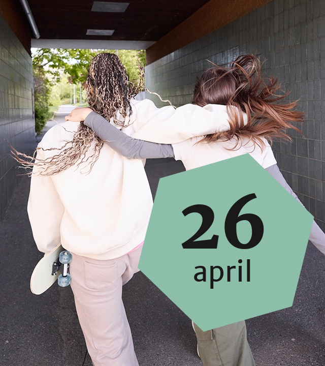 Två tjejer går genom en tunnel. De håller om varandra, och en av dem håller i en skateboard. Vi ser dem bakifrån. Ovanpå fotot ligger en grön sexkantig platta med texten: 26 april.