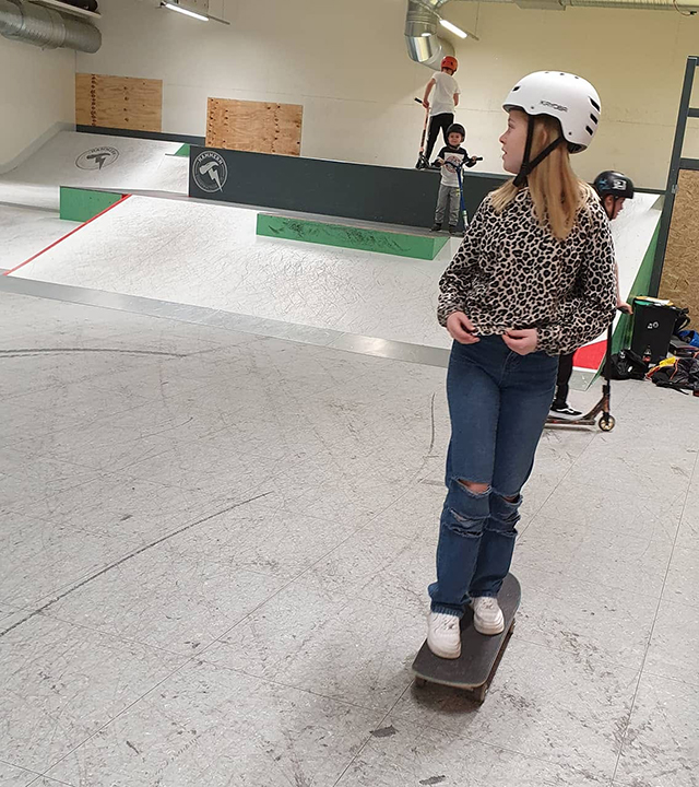 En flicka i tioårsåldern står på en skateboard. I bakgrunden syns barn som står på sparkcyklar. 