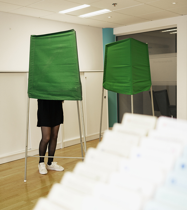 I en vallokal syns två gröna bås. I ett av båsen står en person. I förgrunden syns en ställning med röstningslappar. 