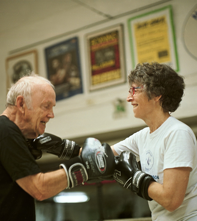 En äldre man och en äldre kvinna boxas med varandra i en träningslokal, iklädda svarta boxningshandskar.