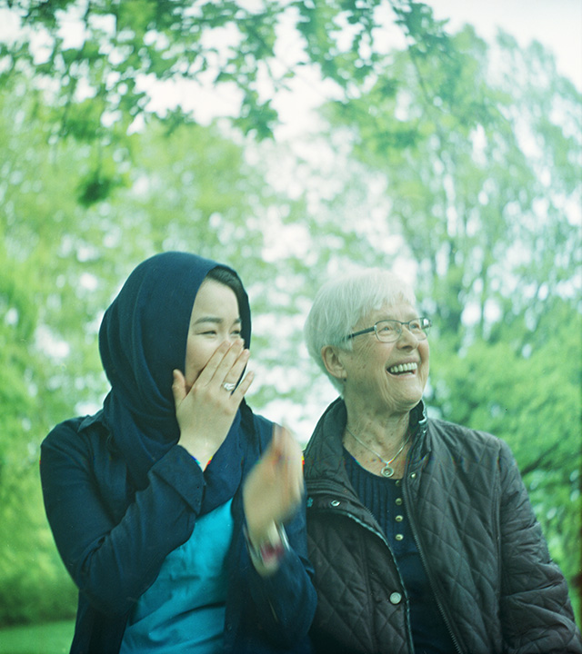 En ung kvinna i slöja och en äldre kvinna i glasögon skrattar tillsammans i gröna omgivningar.