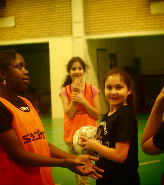 I en idrottshall håller en flicka i en fotboll. Hon är omgiven av tre andra flickor.