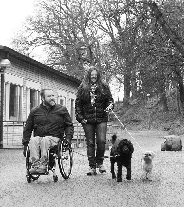 En man i rullstol och en kvinna som går bredvid, båda håller varsin hund i koppel